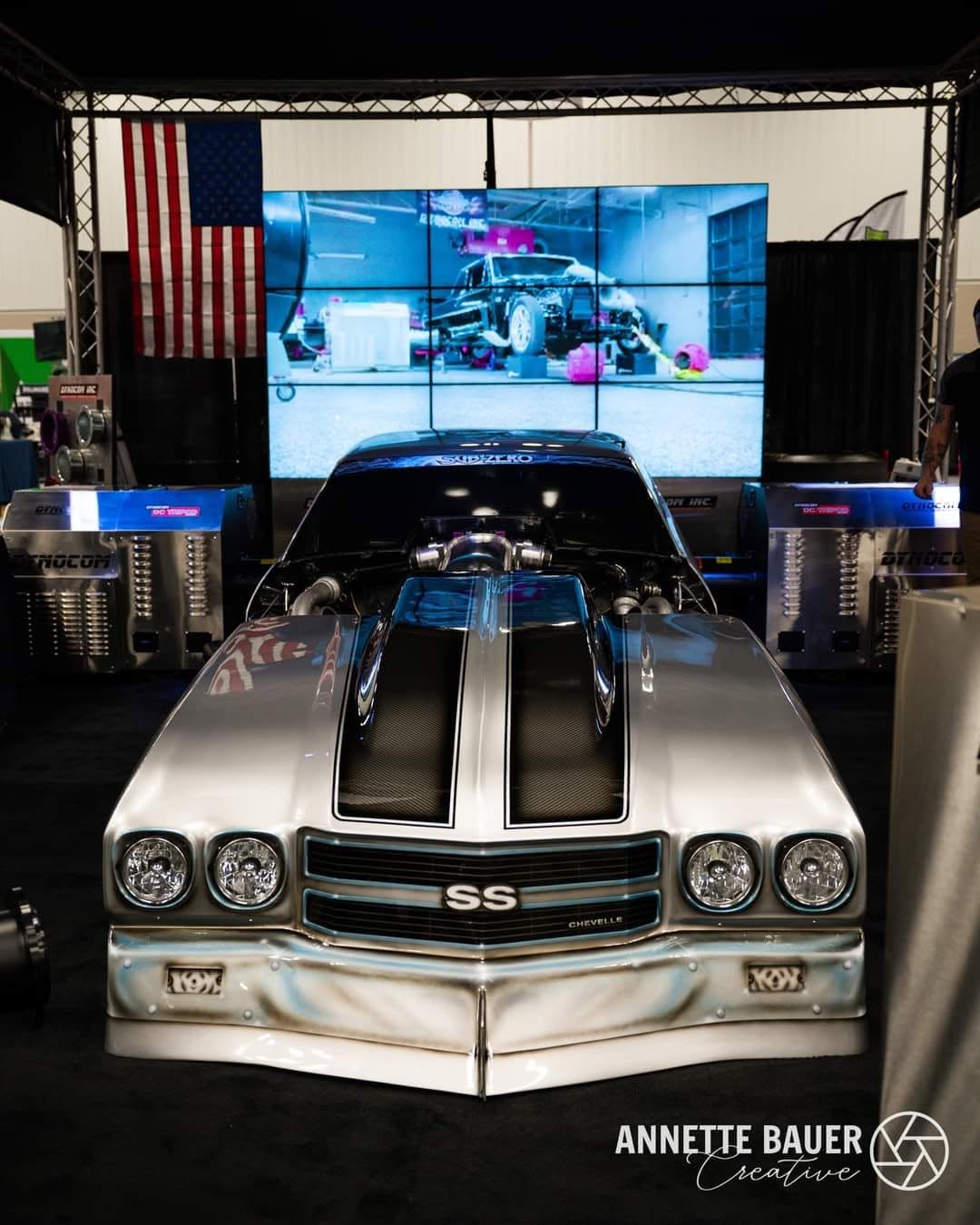 Chevelle SS twin-turbo on Dynocom DC TriPod dynamometer at trade show booth with American flag and video wall