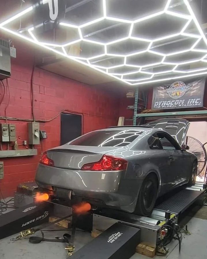 Silver Infiniti G35 spitting exhaust flames on Dynocom AWD dyno under hexagon LED lights at Dynocom Inc shop